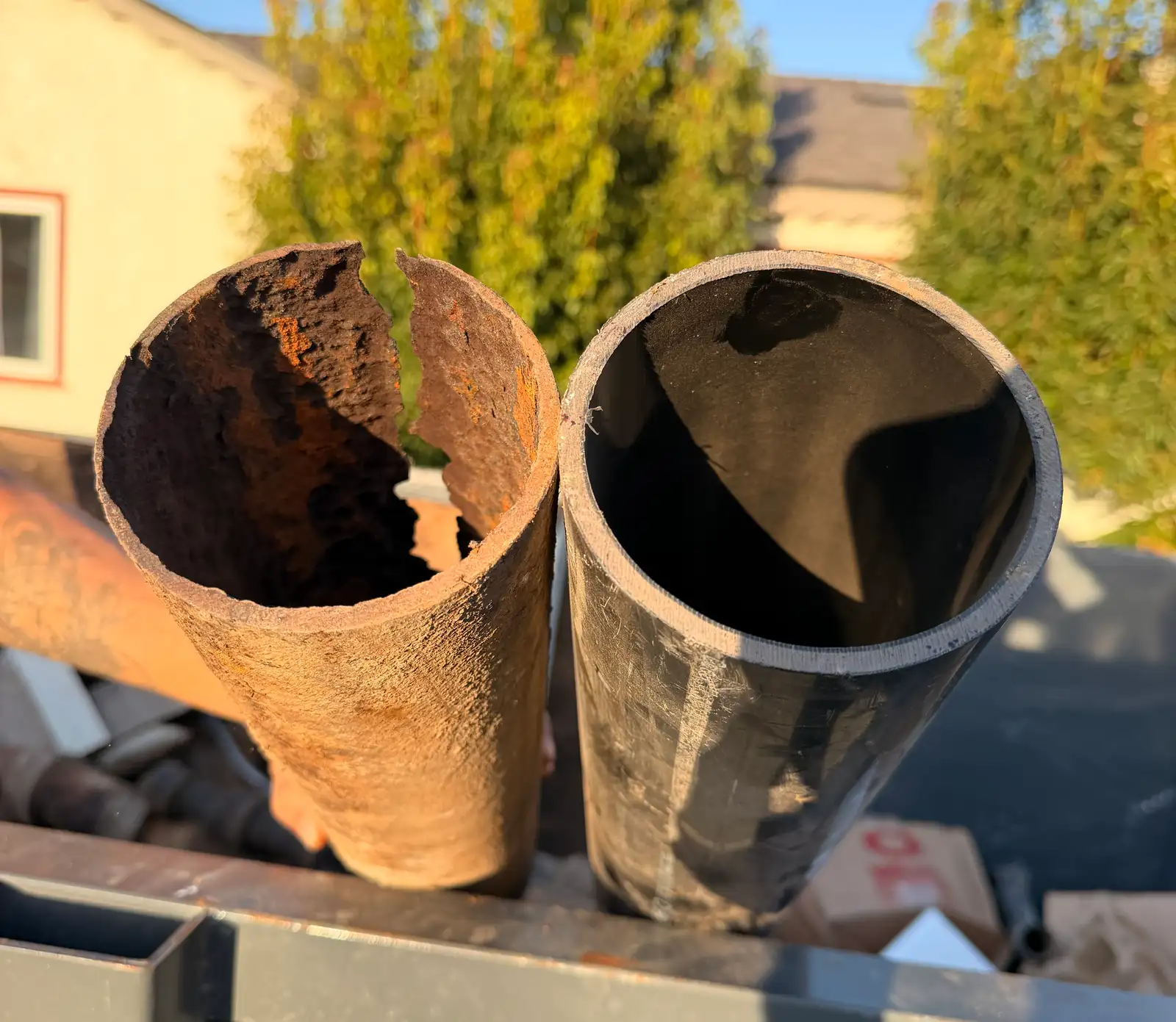 Cut section of failed cast iron sewer pipe held beside a new length of the same nominal diameter on a truck bed, the old piece split open along its length with rust tuberculation closing most of the bore while the new pipe is clean and full width