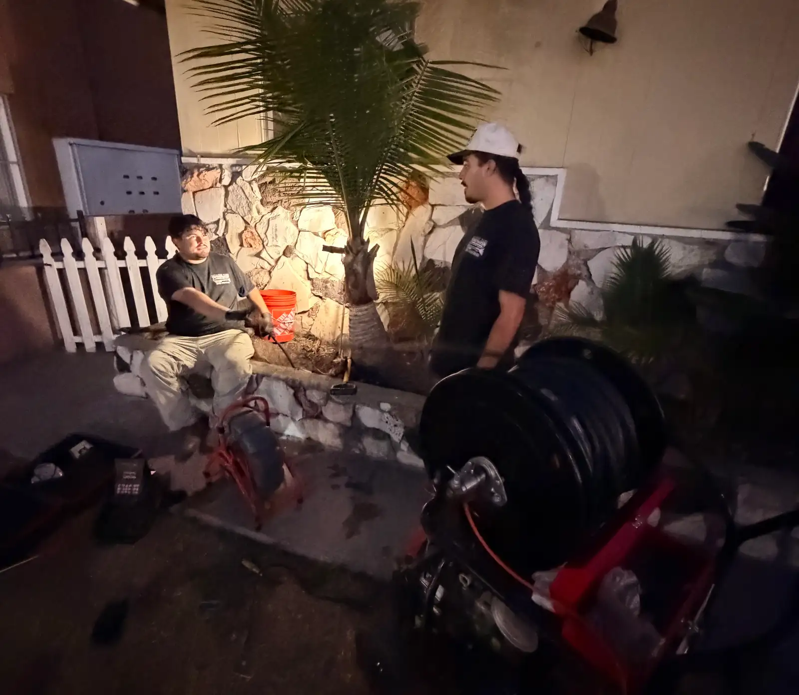 Two Mainline technicians working a residential drain call after dark in Hawthorne, a large drum cable machine staged in the foreground and a second sectional machine on the walkway, the house lit by work light