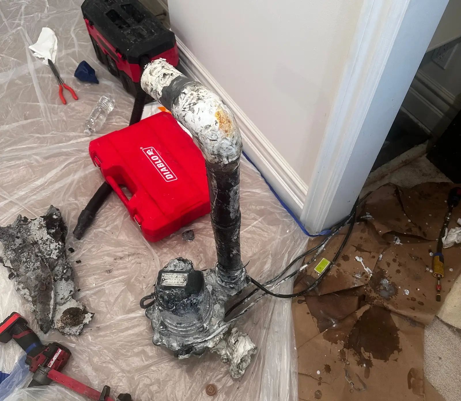 Failed pit pump pulled out and set on plastic sheeting in the finished hallway of a Manhattan Beach home, caked in white mineral scale, with floor protection taped along the baseboard and tools staged on the plastic