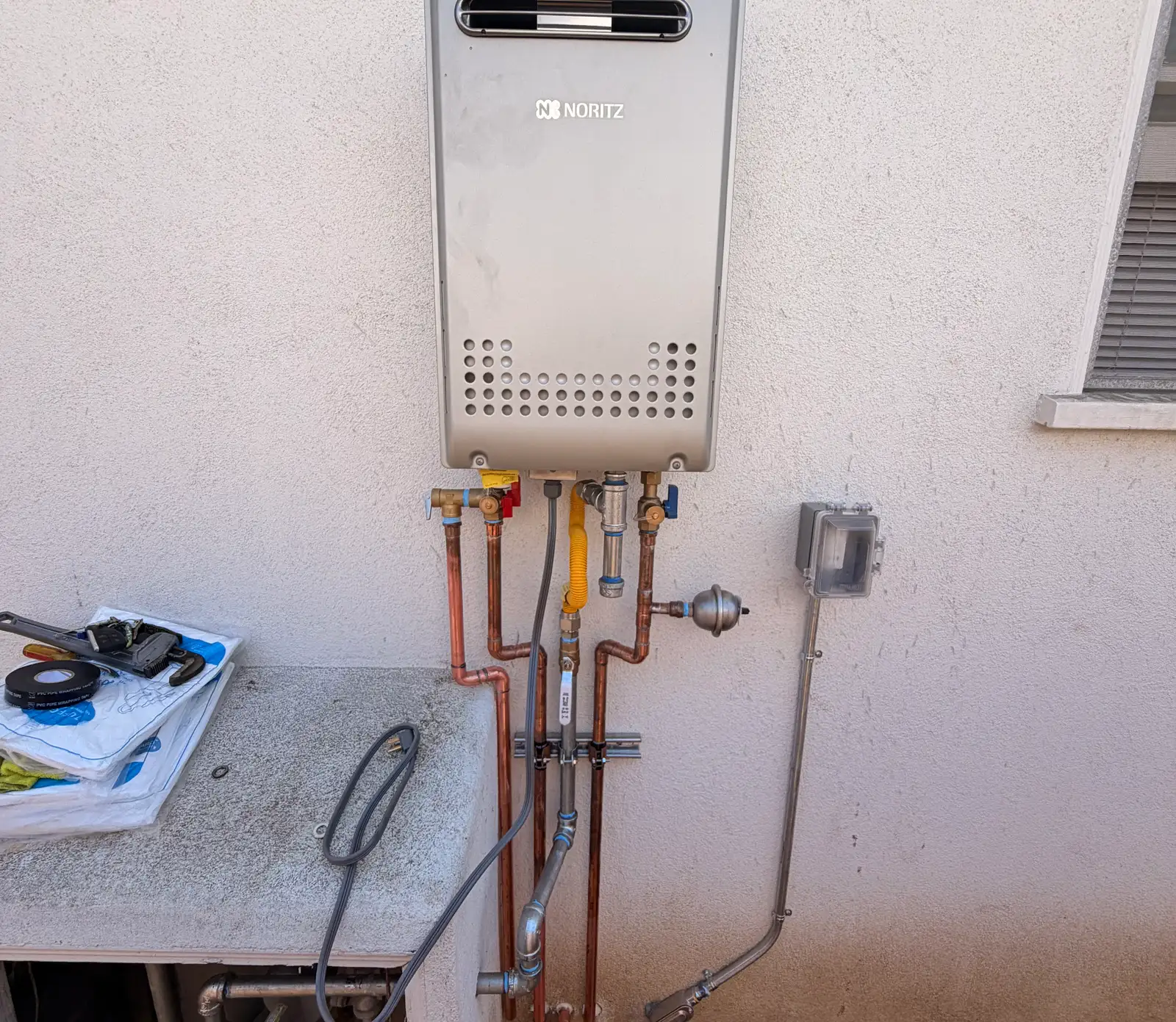 Wall-mounted Noritz tankless water heater installed outdoors on a stucco wall, with copper hot and cold lines, red and blue isolation service valves, a yellow corrugated gas connector with its own shutoff, condensate drain and a weatherproof electrical disconnect alongside