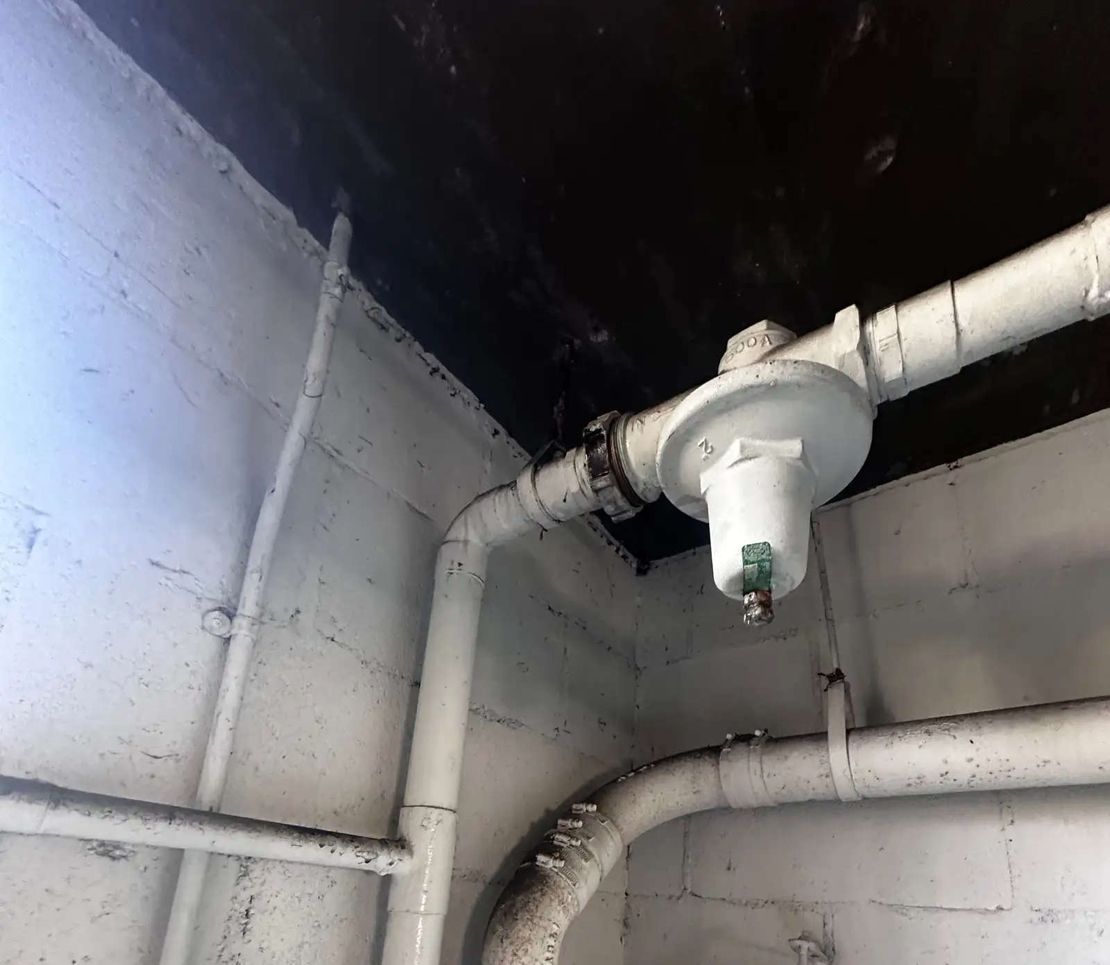 Old pressure reducing valve on a 2-inch building water main, painted over white along with the surrounding piping and block wall, with a green adjustment tag and a black-handled gate valve nearby, before replacement