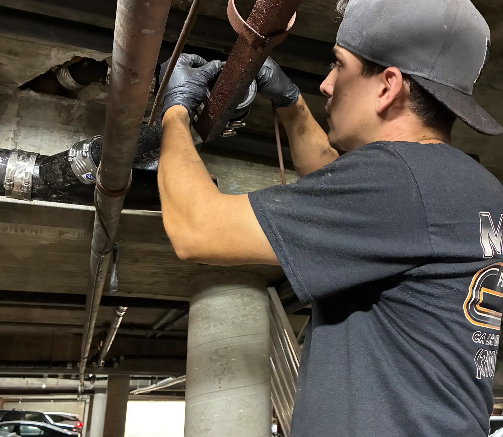 Mainline technician in a branded shirt cutting into corroded overhead cast iron drain pipe on the underside of a subterranean parking deck, with shielded no-hub couplings already fitted, parked cars and caution tape visible behind him
