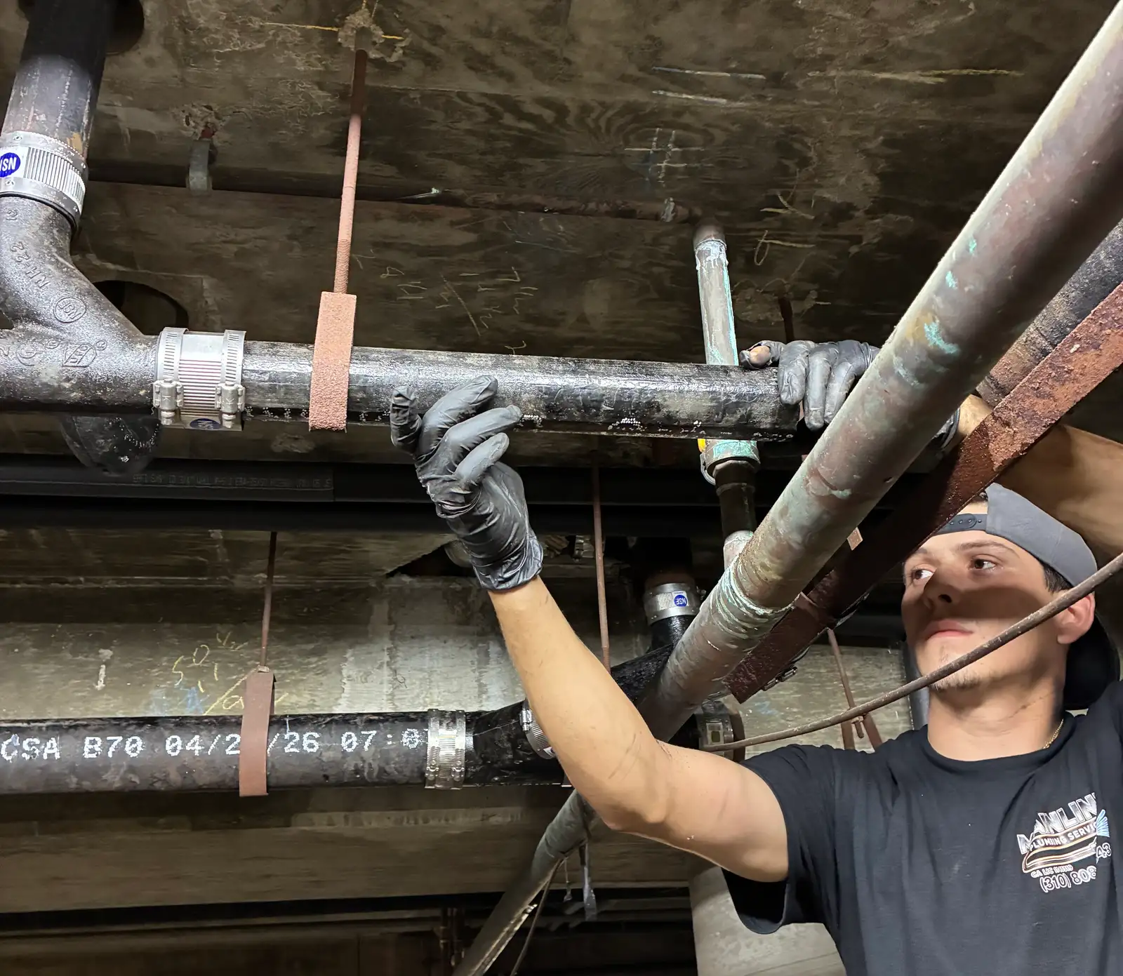 Newly installed no-hub cast iron drain line hung beneath a parking structure deck with shielded band couplings and fresh strap hangers, the pipe carrying a legible CSA B70 mill stamp dated 2026