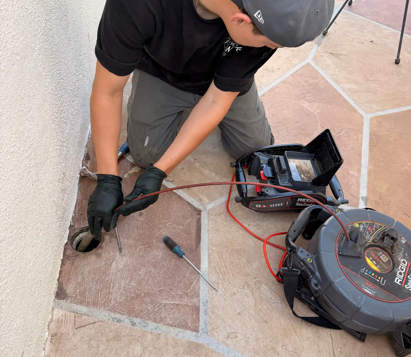 Mainline technician feeding a sewer inspection camera into a flush cleanout port in a stamped concrete patio, with the camera reel and monitor beside him and nothing dug up