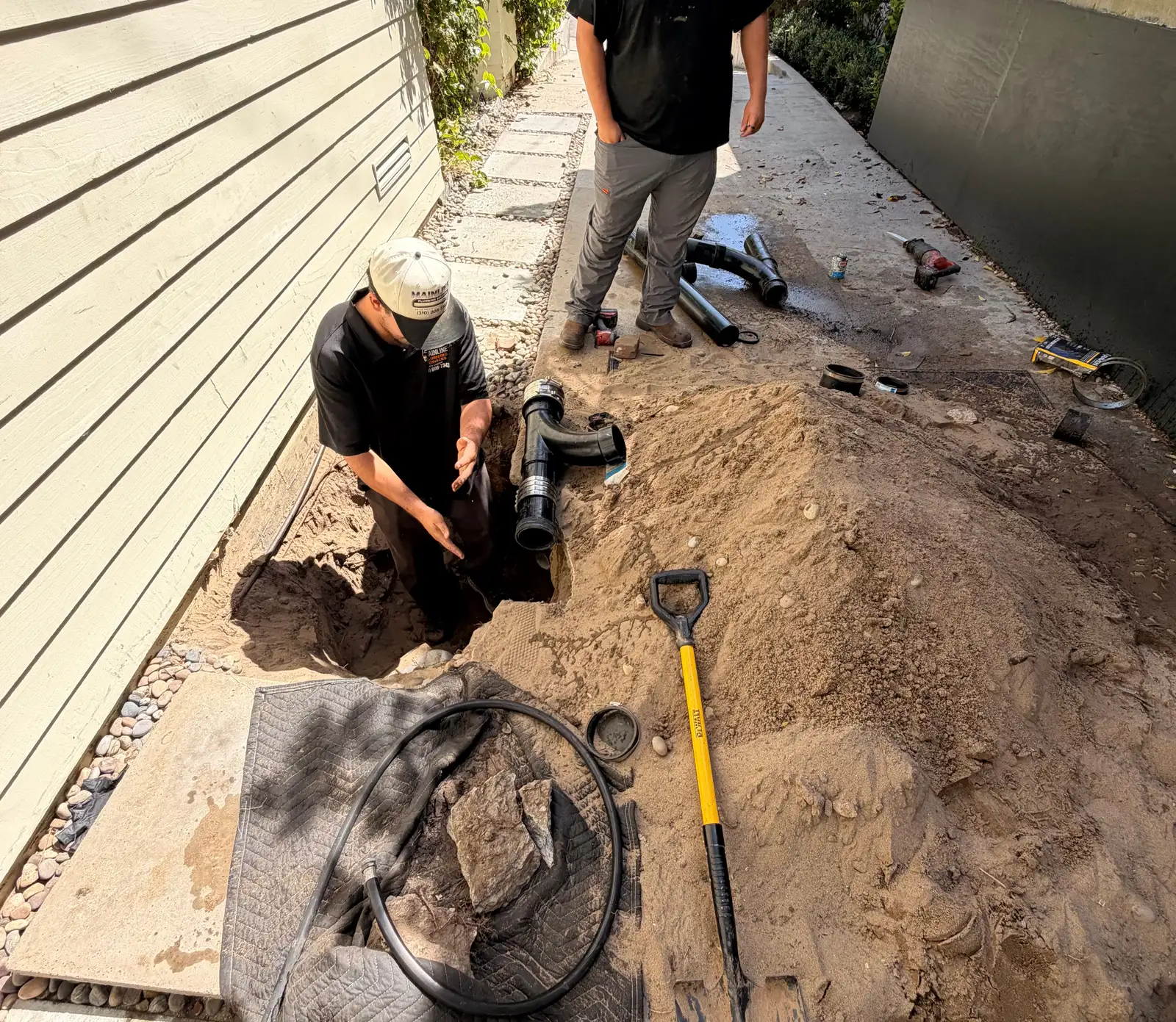 Mainline crew member kneeling in one small access pit beside a house, with a new black ABS cleanout wye and shielded couplings ready beside the pit and the dug soil piled along the side-yard walkway