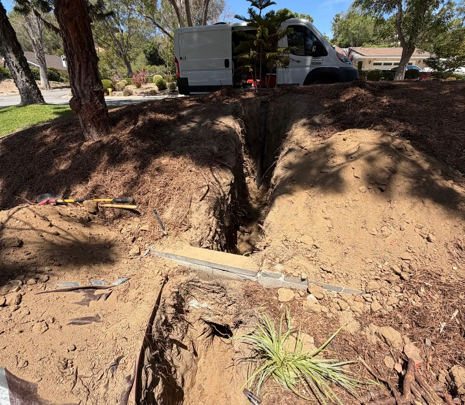 Open trench excavation for a sewer lateral replacement cutting through a landscaped front yard, with spoil piled either side, a walkway cut through and mature tree roots severed