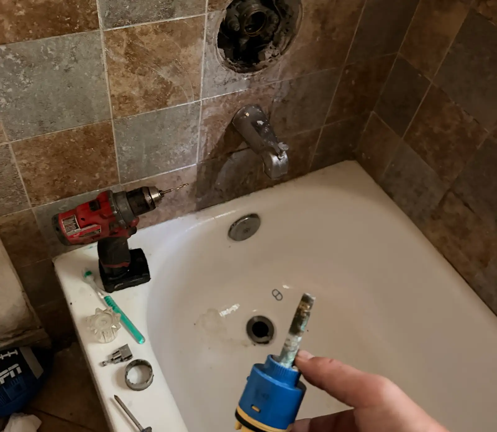 Shower and tub valve cartridge just removed and held to camera, its stem and seals furred with white mineral scale, with the open valve body in the tiled wall and the tub spout behind it