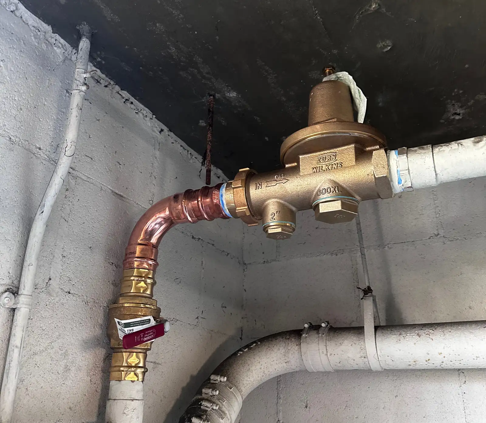 New 2-inch Zurn Wilkins 600XL pressure reducing valve installed on a building water main in a block-wall utility room, fed by new copper with a ball valve upstream, beside the original painted piping